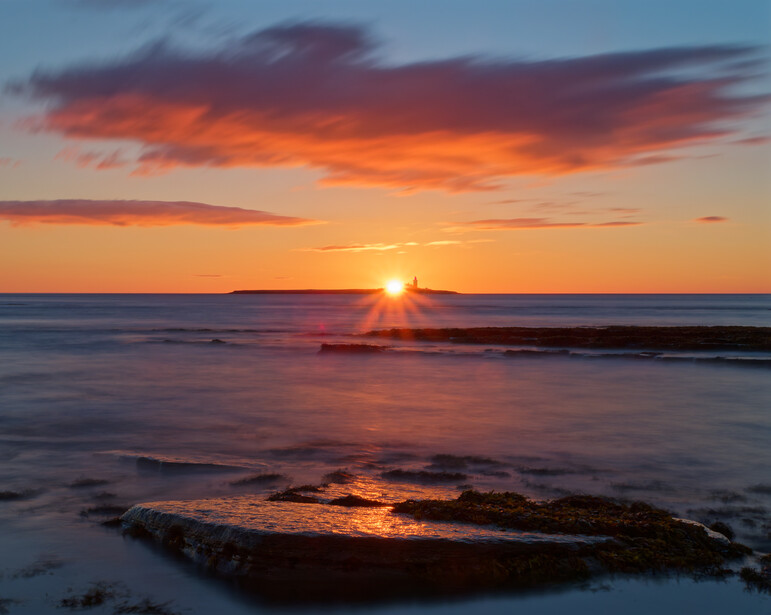 Sun breaking through orange clouds above the ocean horizon at sunset, with rocky outcroppings in the foreground.