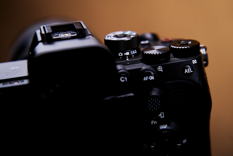 Close-up of a mirrorless camera body showing control dials, buttons, and flash unit against warm background.