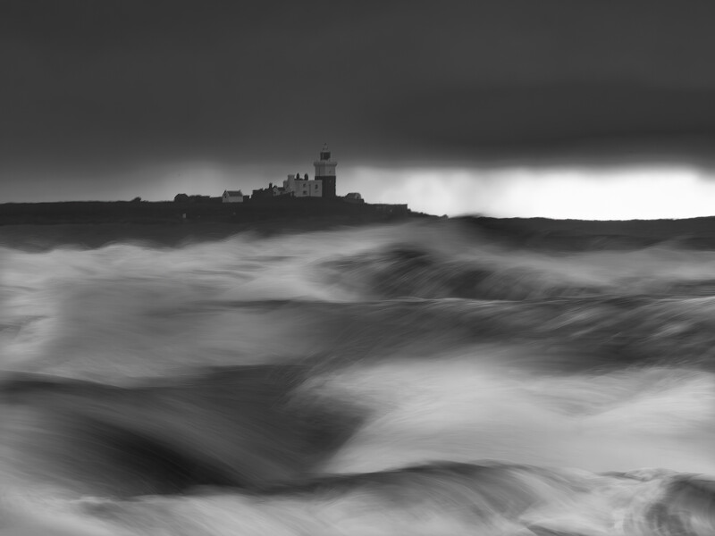 Silhouetted castle on a rocky headland surrounded by turbulent, motion-blurred waves under dark skies.