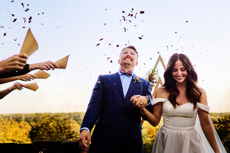 Couple walking arm-in-arm outdoors with confetti falling and golden paper cones being held above them.