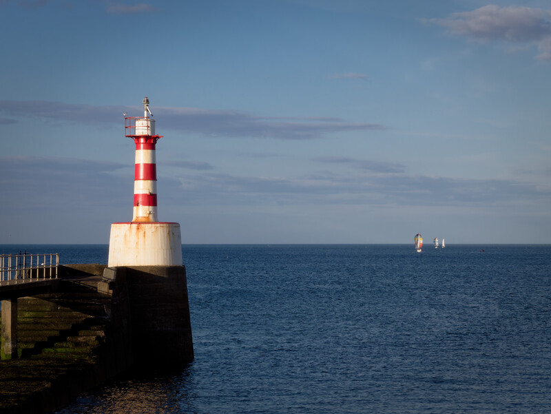 Red and white striped lighthouse on a stone pier extending into calm blue sea.