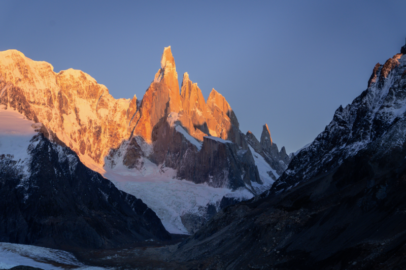 Snow-covered mountain peaks glowing orange in alpenglow at dawn, with a glacier visible in the valley below.