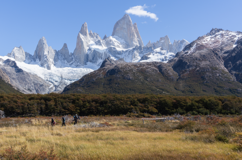 Snow-capped granite peaks rising behind a golden grassland with hikers and forest in the middle distance.