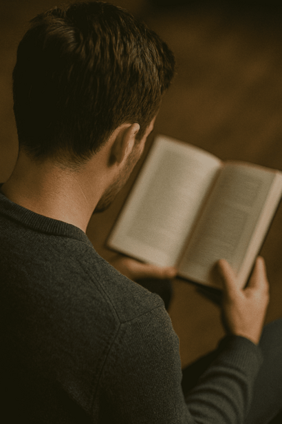 Person reading an open book while seated, photographed from behind in warm, dimly lit indoor setting.