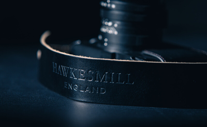 Close-up of a black leather camera strap with embossed text reading "HAWKESMILL, ENGLAND" against a dark blue background.