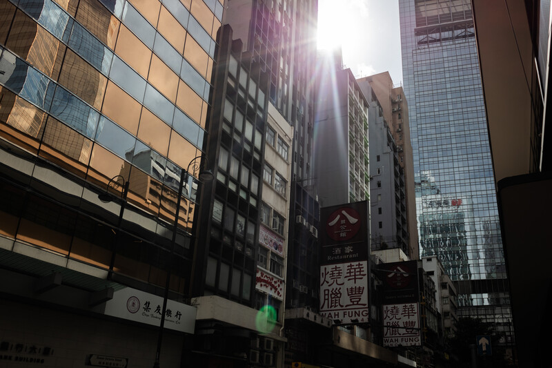 Urban street canyon with mixed-height buildings backlit by bright sun, casting deep shadows and creating strong contrast.