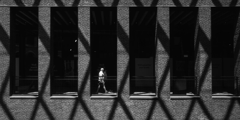 Solitary figure walking through geometric shadow patterns cast on a brick building facade.