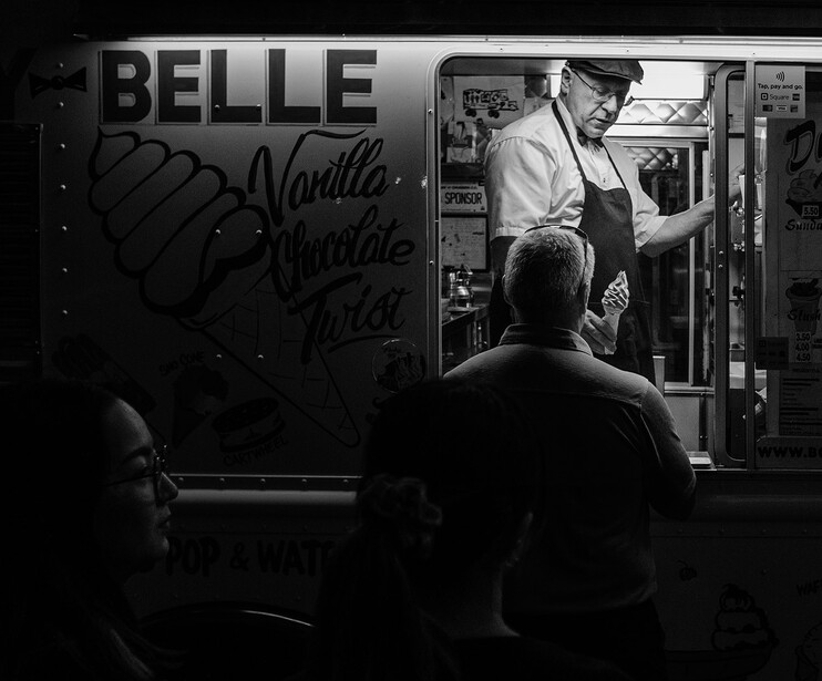 Black and white street photograph of a vendor serving a customer through a food service window at night.