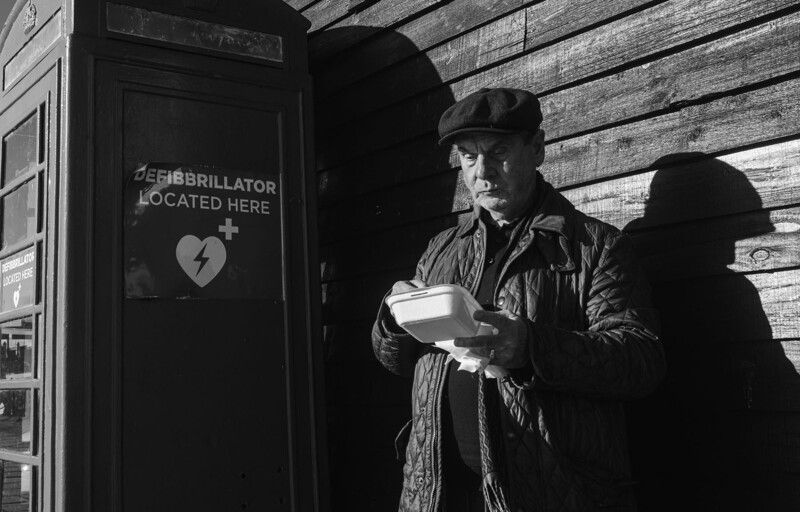 Two figures in dark clothing stand near a defibrillator cabinet mounted on a wooden wall in black and white.