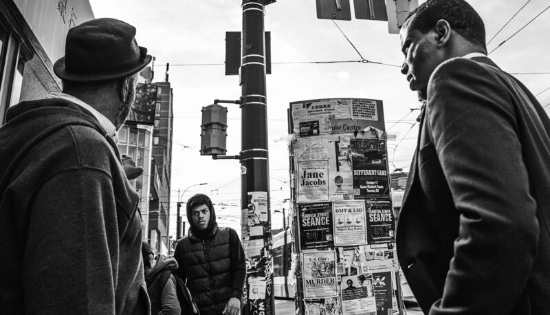 Three people on an urban street with posted advertisements covering a utility pole behind them.