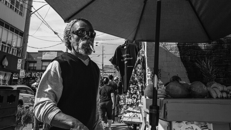 Man wearing sunglasses and vest at an outdoor market stall under a black umbrella.
