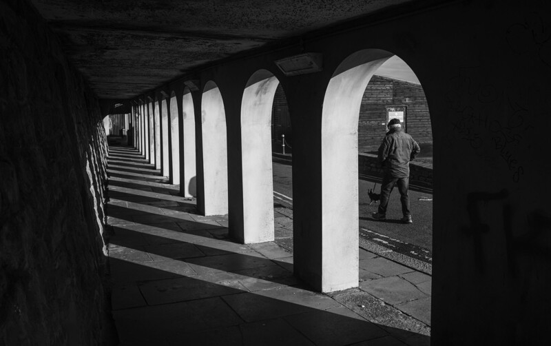 Repeating white concrete columns receding into shadow beneath a dark overhead structure, with a solitary figure visible through an arch in the distance.