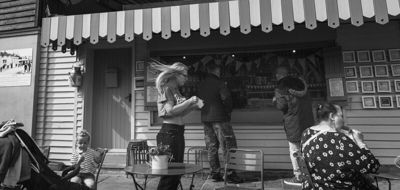Black and white street photography showing several people interacting outside a storefront with an awning.