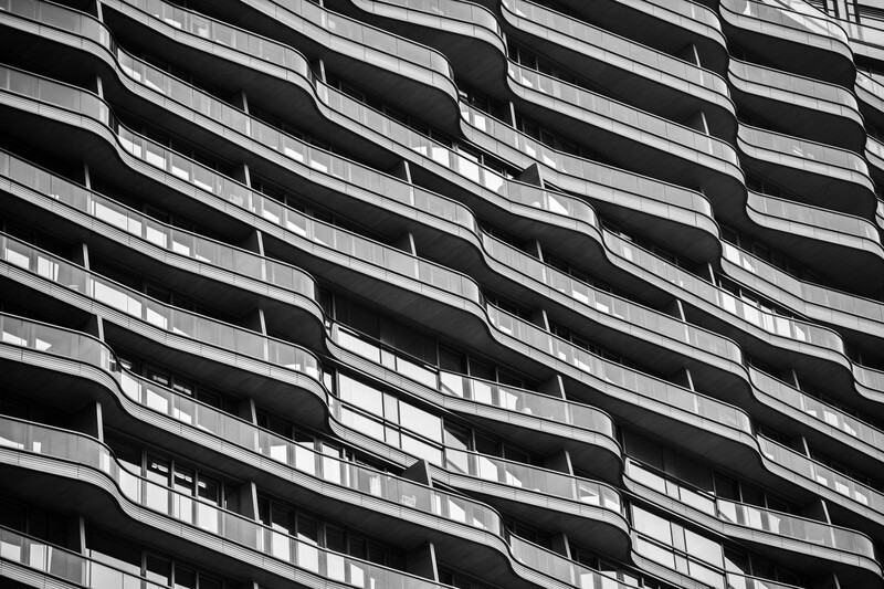 Black and white photograph of a modern building facade with curved, stacked balconies creating dynamic horizontal lines and deep shadows.