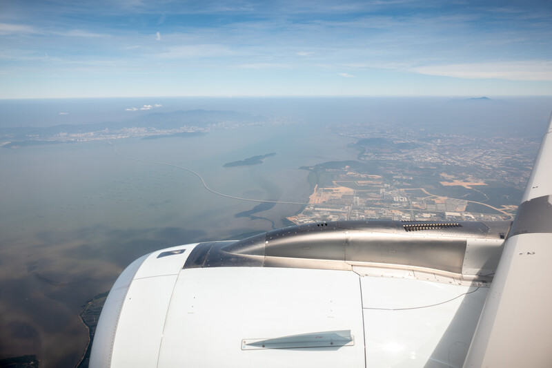 Aerial view from aircraft window showing wing, engine, and landscape with water and industrial facilities below.