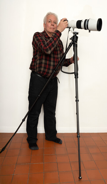 Older man in plaid shirt looking through large telephoto lens mounted on tripod.