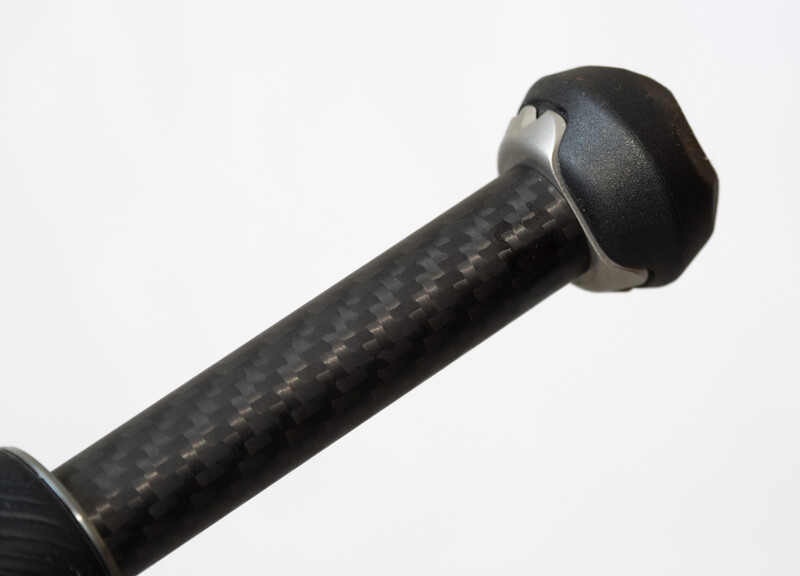 Close-up of a camera tripod head with textured black carbon fiber pole and metal ball mount against white background.