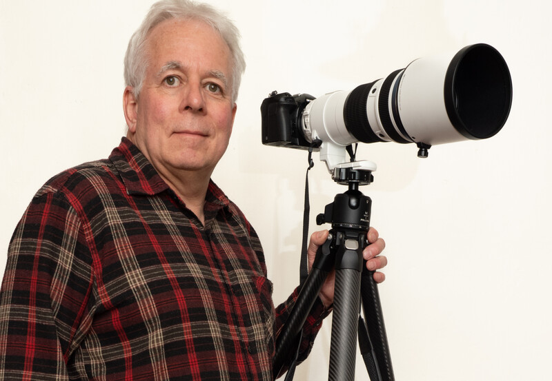 Photographer holding a telephoto lens mounted on a tripod with a carbon fiber leg.