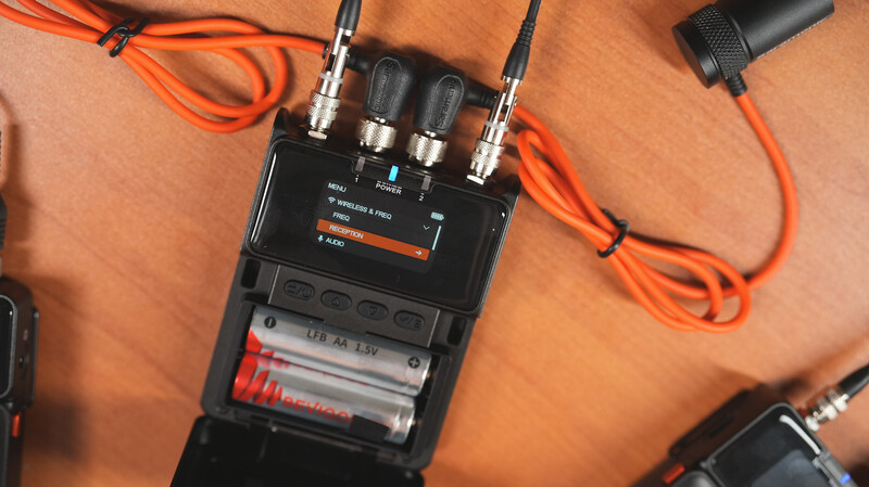 Wireless audio receiver with four XLR connectors, orange cables, and battery compartment on orange fabric backdrop.