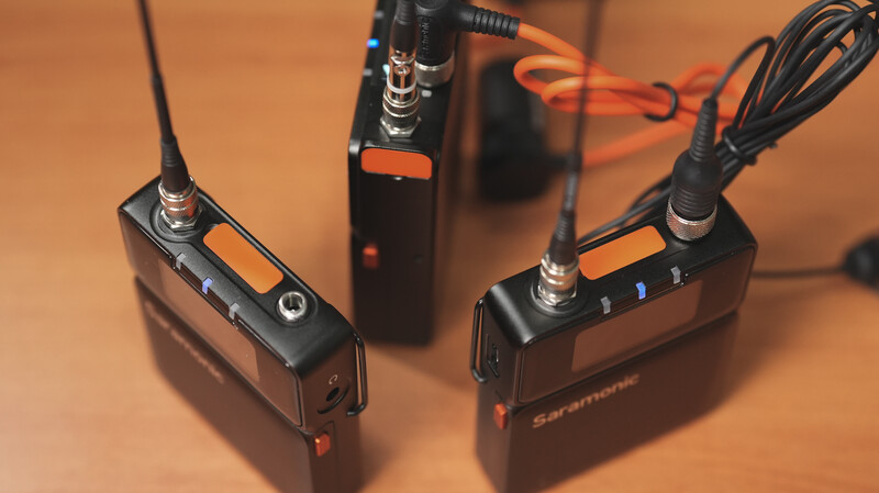Three wireless lavalier microphone transmitters with antennas arranged on a warm orange surface.