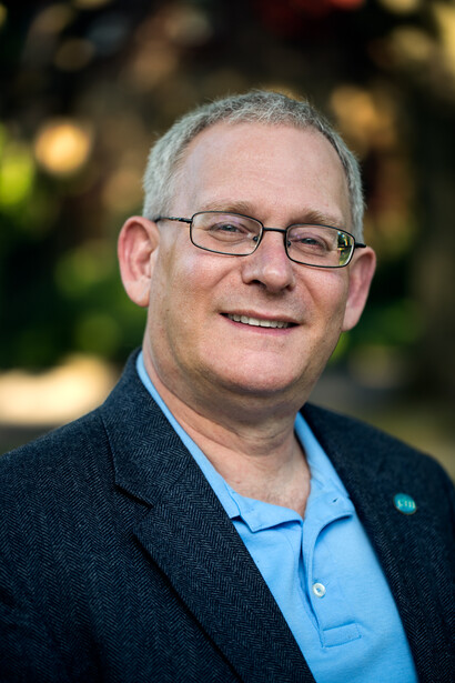 Man wearing glasses and blue polo shirt smiling at camera in outdoor setting.