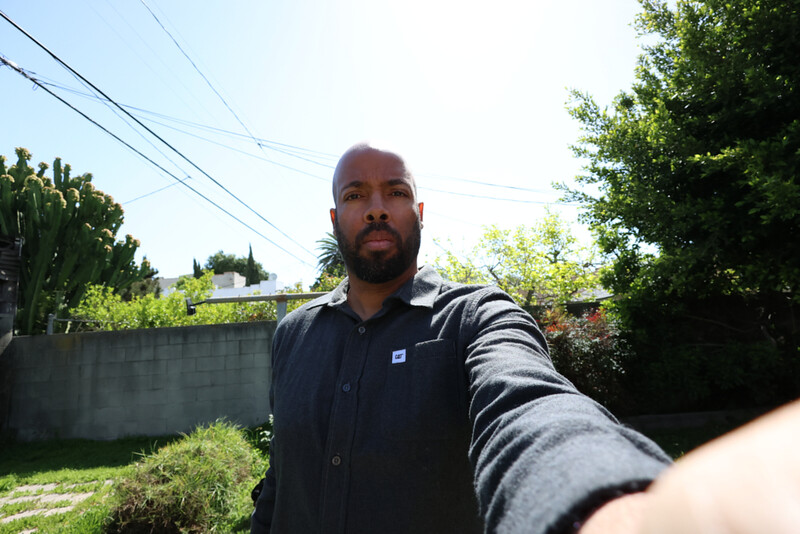 Man taking a selfie outdoors in natural daylight with residential setting in background.