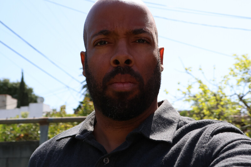 Portrait of a man with a full beard, looking directly at the camera with a neutral expression outdoors.