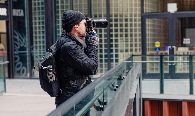 Photographer with backpack and telephoto lens standing on an urban pedestrian bridge, framing a shot.