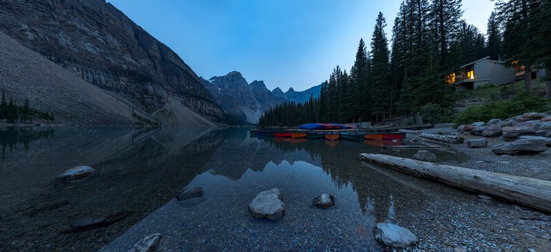 Alpine lake at dusk with moored canoes, rocky shoreline, forested mountains, and illuminated lodge.