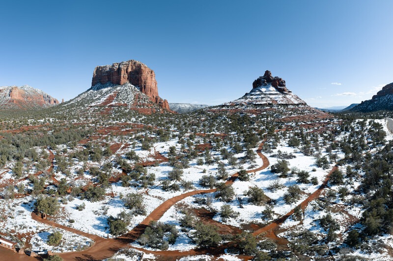 Snow-covered desert landscape with distinctive red rock buttes and scattered evergreen trees under clear blue sky.