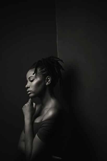 Profile of a woman against a dark wall, hand raised to her neck, in dramatic black and white lighting.