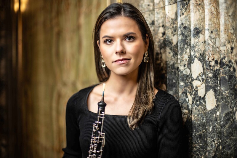 Woman holding an oboe, posed against a weathered stone wall.
