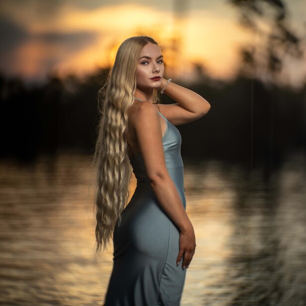 Woman with long blonde hair posing by water at sunset, wearing a fitted sleeveless dress.