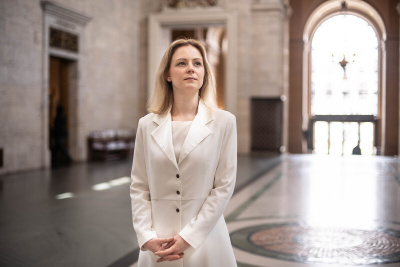 Professional woman in cream blazer standing in classical hallway with arched doorway and natural light.