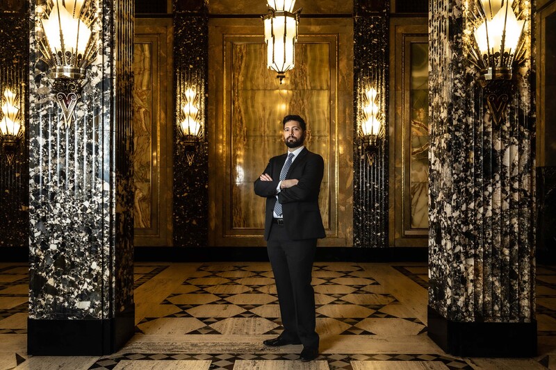 Man in dark suit standing in ornate hallway with symmetrical columns and gold-toned décor.