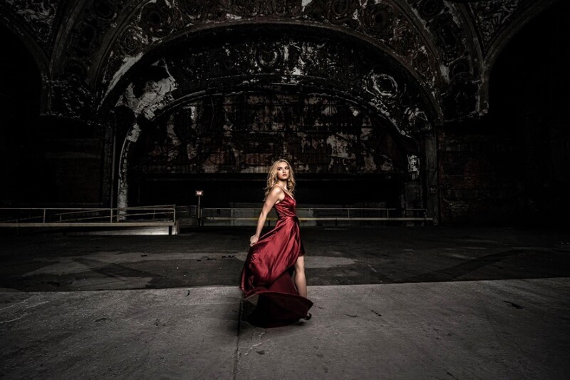 Woman in burgundy gown kneeling on concrete floor facing abandoned stage structure.