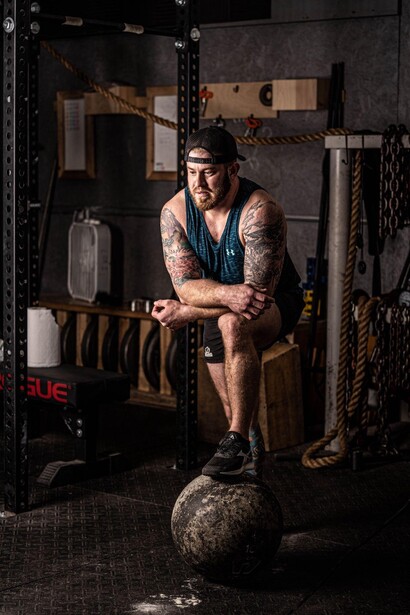 Tattooed man in tank top sitting on medicine ball in industrial gym setting.
