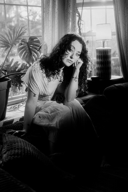 Woman with curly dark hair resting her head on her hand, sitting by a window in natural light.