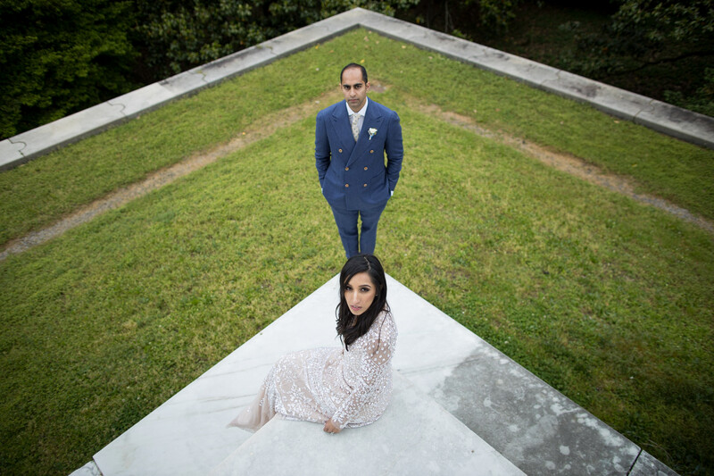 Bride in lace dress and groom in navy suit standing on white concrete path on green rooftop, photographed from above.