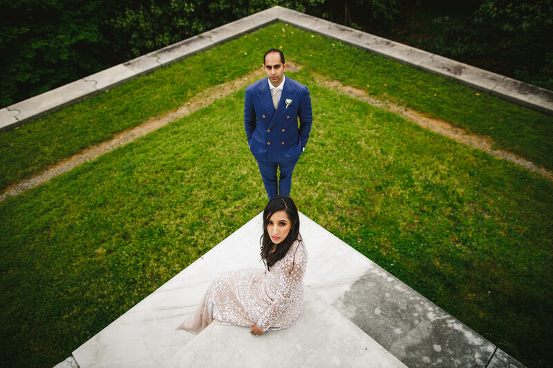 Bride in lace dress and groom in blue suit standing on concrete path through green lawn, photographed from above.