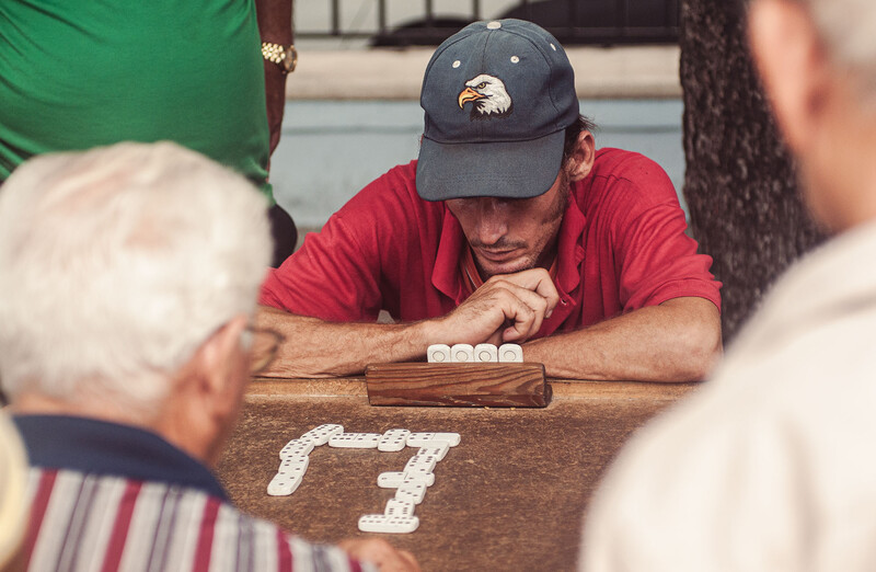 Man in red shirt and dark cap concentrating on domino tiles arranged on wooden surface during outdoor game.
