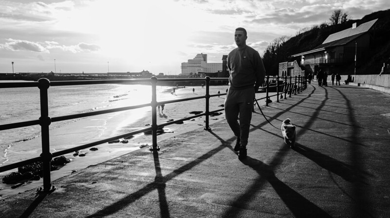 Person in wetsuit walking along a waterfront promenade with a small dog, long shadows cast across the pavement.
