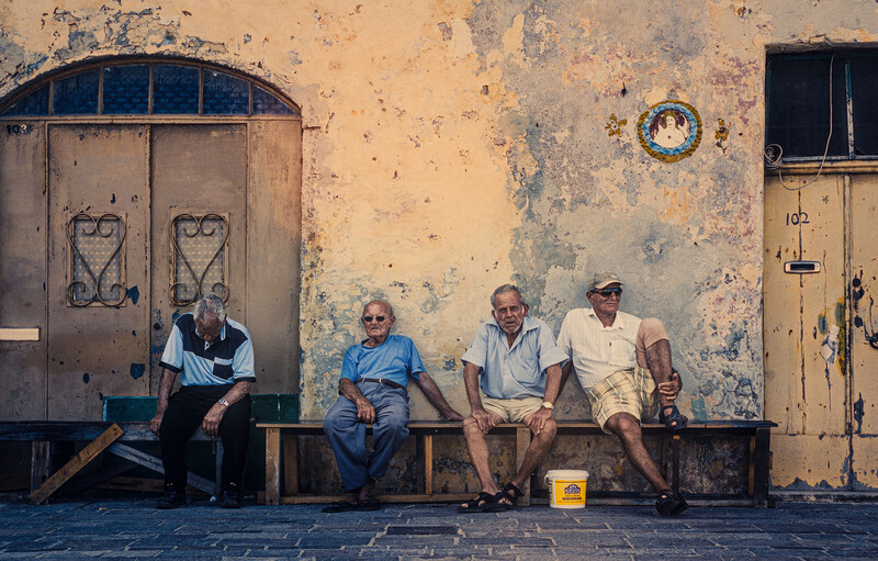 Four elderly men sitting on a bench against a weathered yellow wall with ornate doors.