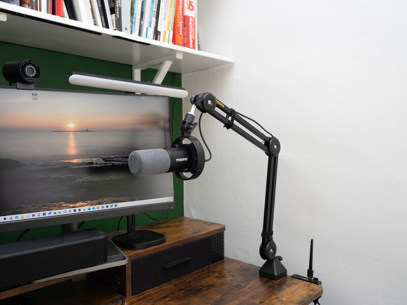 Professional microphone mounted on articulating arm above wooden desk with monitor displaying sunset landscape image.