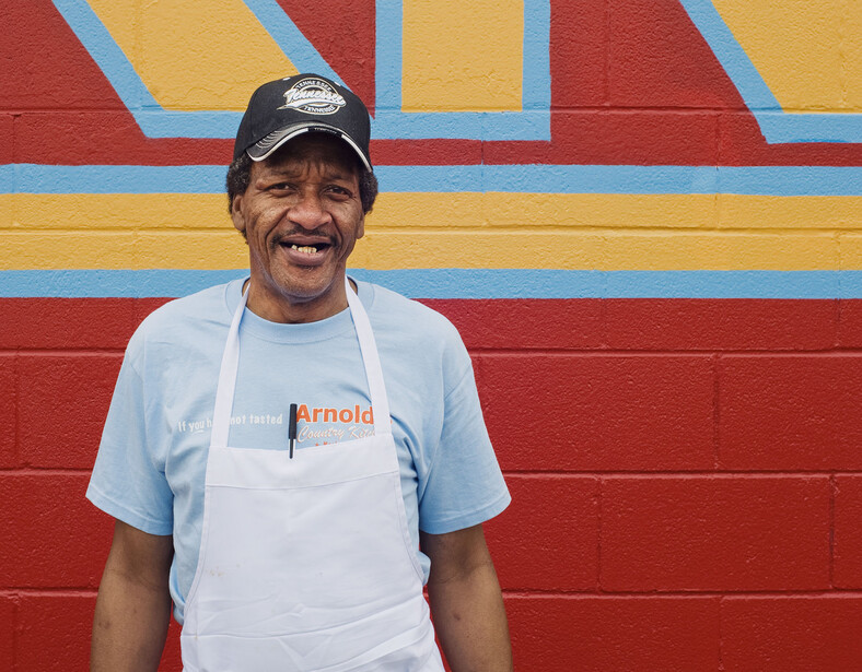 Man wearing light blue apron and cap smiling in front of colorful striped wall backdrop.