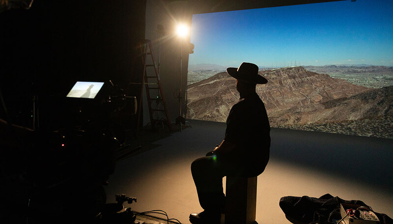 Silhouetted figure in cowboy hat sitting on stool while viewing large landscape photograph lit by studio lighting.
