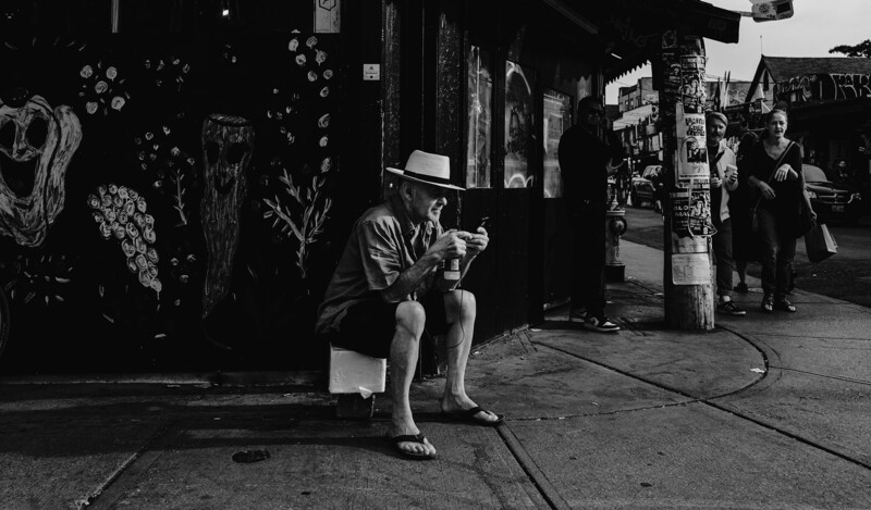 Person sitting on curb in urban alley, wearing fedora and light-colored shorts, reading printed material.