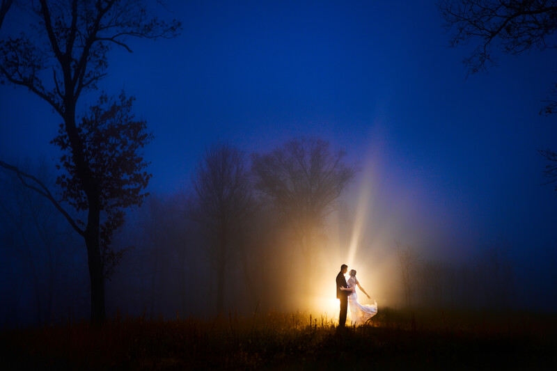 Silhouetted couple standing in a field at dusk, illuminated by golden backlighting creating a bright halo effect.