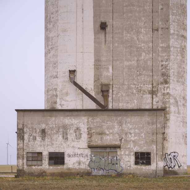 Weathered concrete grain elevator with deteriorating facade and metal chute against overcast sky.