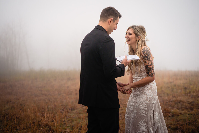 Groom in black suit exchanges vows with bride in lace dress in misty field.